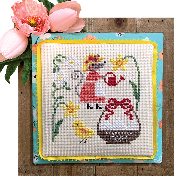 Cross-stitched picture of a woman with a chick and flowers on a wooden surface.