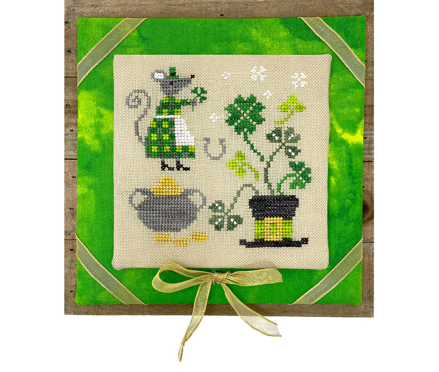 Cross-stitched piece with a cat, teapot, and plants on a green background with a ribbon.