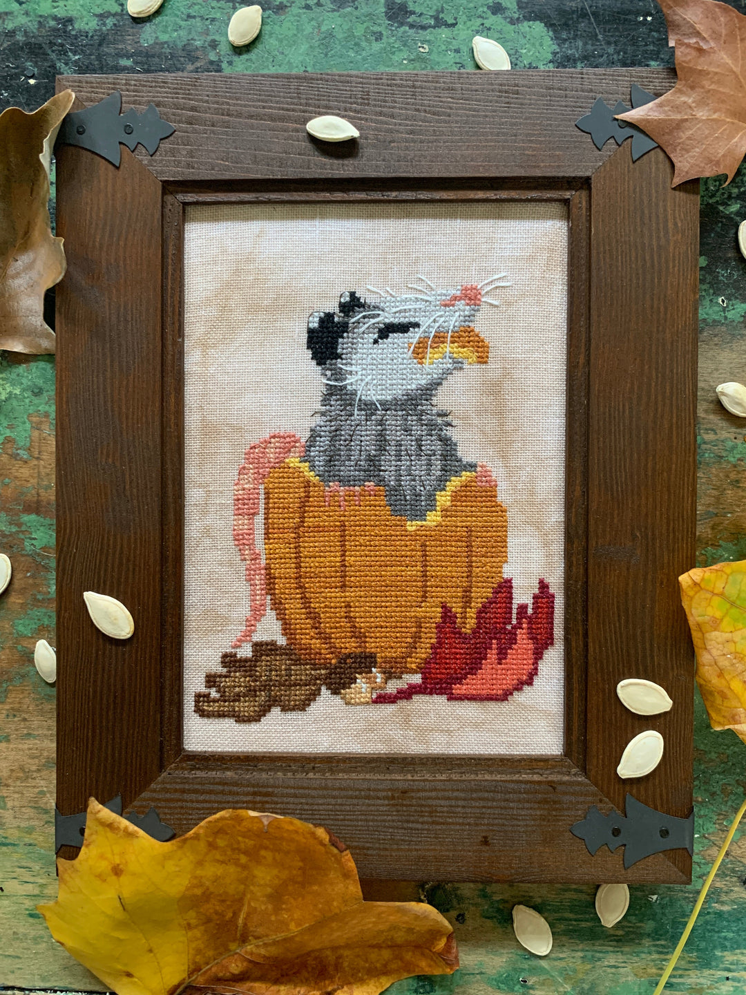 Cross-stitched picture of a bird in a pumpkin on a wooden frame with autumn leaves.