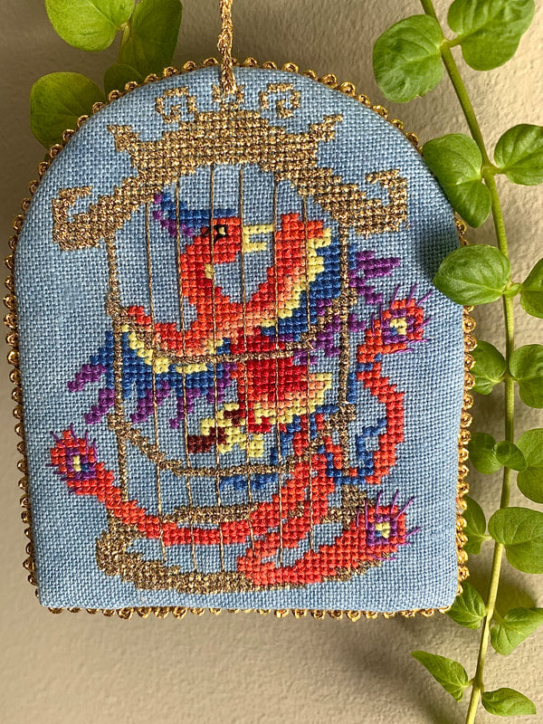 Embroidered artwork of a colorful bird in a cage on a light blue background, surrounded by green leaves.