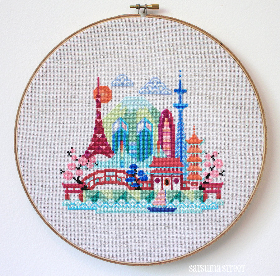 A needlecraft kit depicting a stylized Tokyo skyline, featuring various colorful buildings and a bridge, stitched on a piece of fabric.