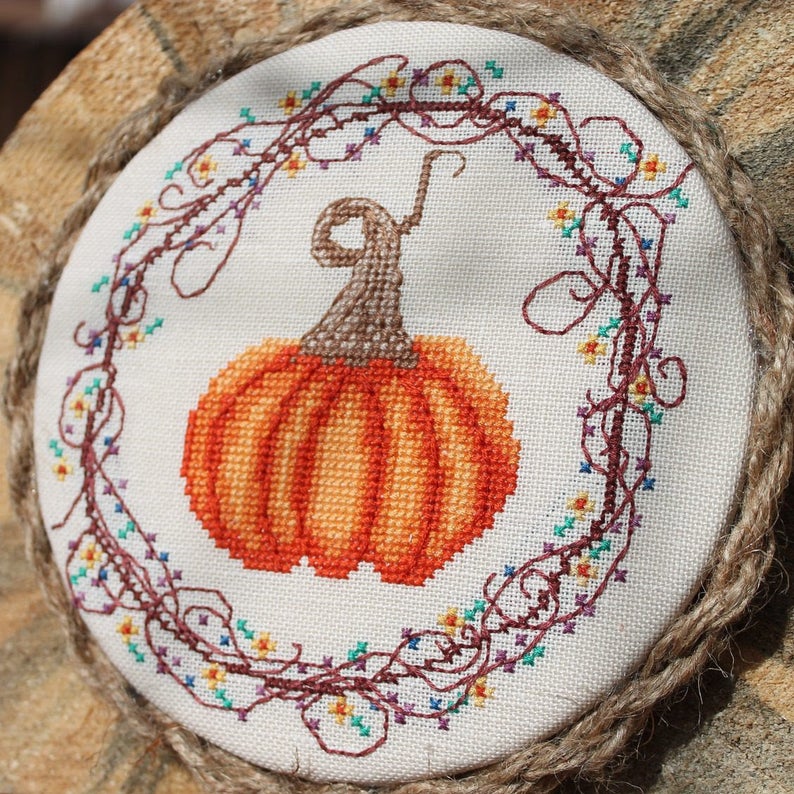 A stitched pumpkin and wreath design in rich Autumn colors on a textured fabric background.