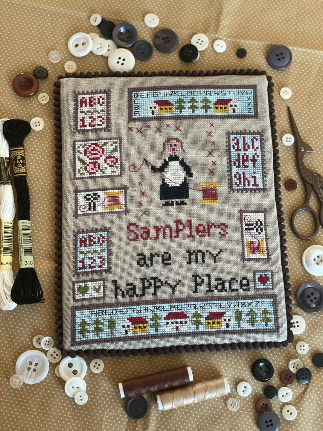 Cross-stitched sampler with text 'Samplers are my happy place' surrounded by sewing materials on a beige background.