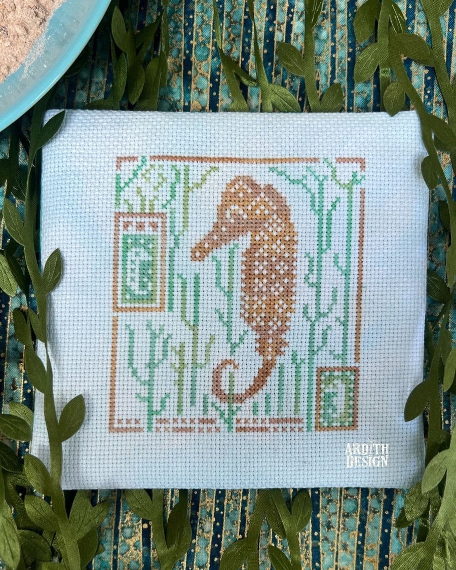 Cross-stitch needlework depicting a seahorse surrounded by aquatic plants, displayed on a blue and white striped surface.