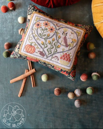 A cross-stitch kit depicting an autumnal scene with hedgehogs, owls, and seasonal plants, framed in a heart-shaped hoop. Various craft materials like floss and pins are also visible.