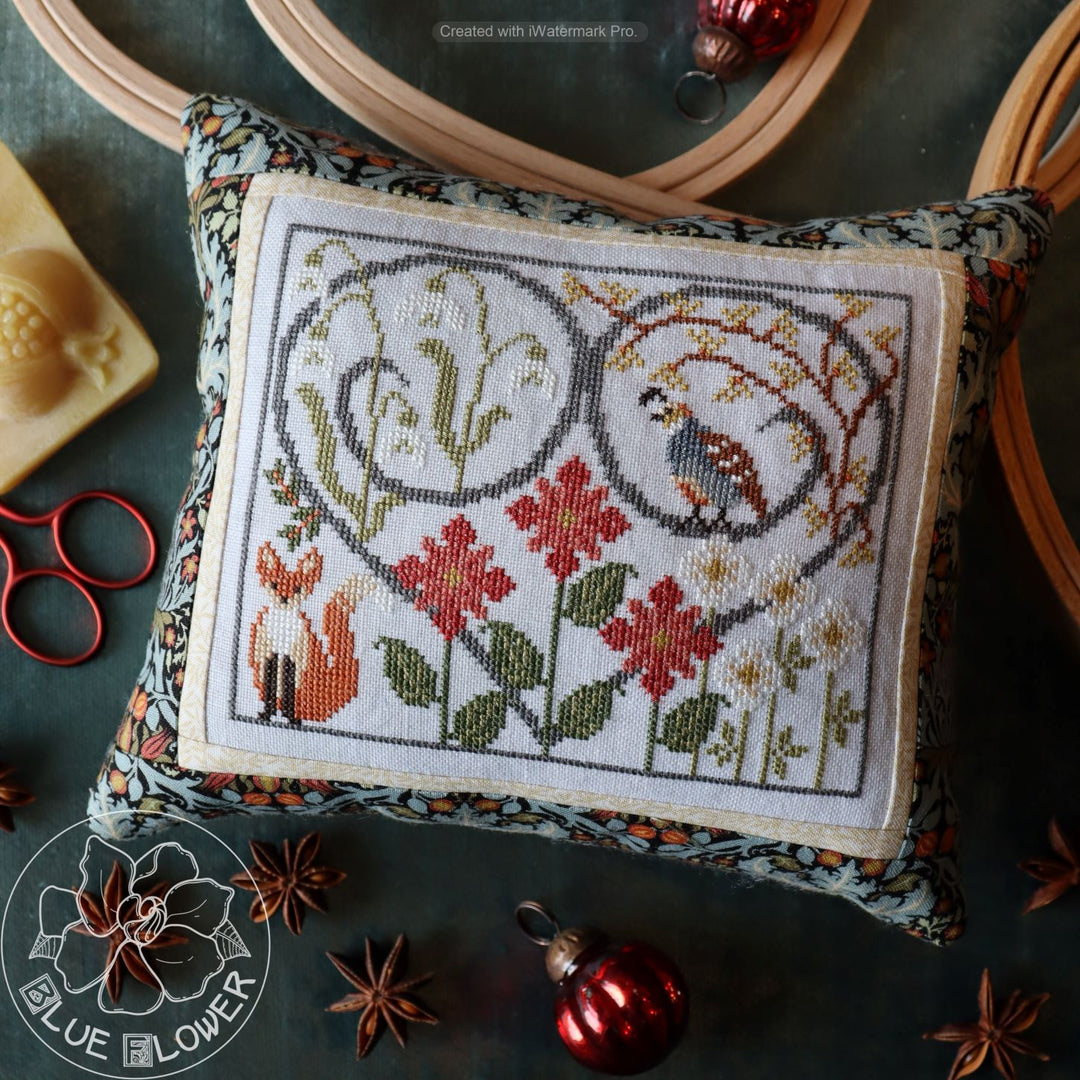 Cross-stitch needlework depicting a winter scene with a fox, quail, snowdrops, and poinsettias, surrounded by Christmas decorations.