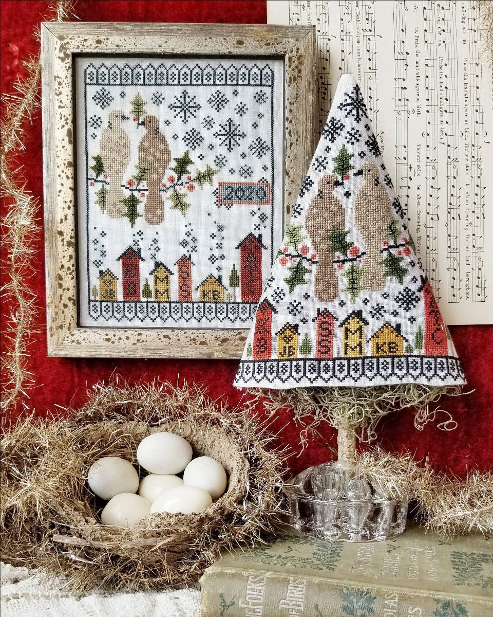 A cross-stitching kit depicting a Christmas scene with houses, trees, and birds, framed and displayed alongside a conical hat with a similar design.