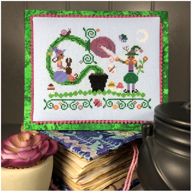 Cross-stitched design with green border on a wooden surface with a cupcake and teapot.