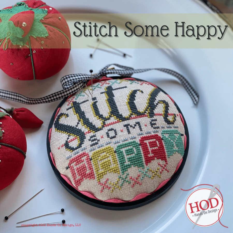 Embroidery hoop with 'Stitch Some Happy' text and colorful cross-stitch design on a white surface.