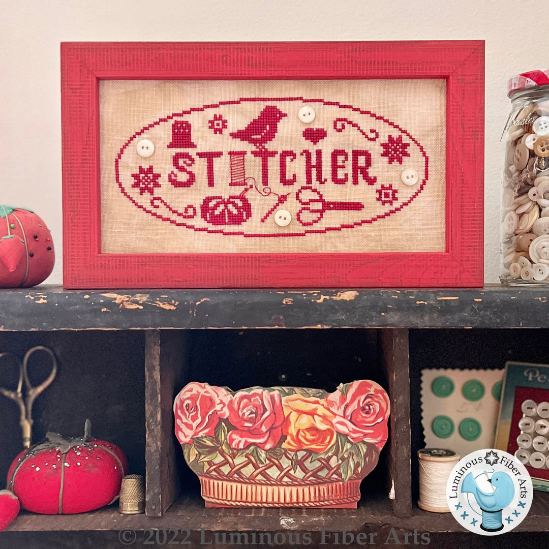 Framed cross-stitched piece with 'Stitcher' text on a shelf with decorative items.