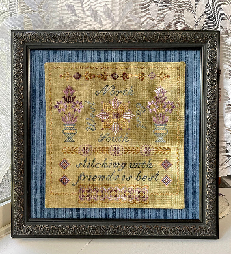 Framed cross-stitched sampler with decorative elements on a textured wall background
