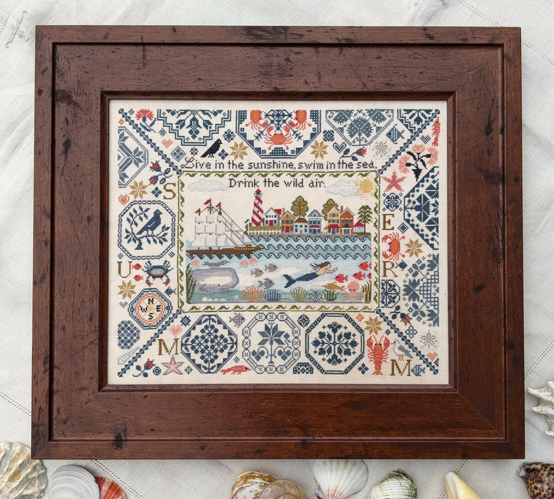 Framed cross-stitched sampler with coastal scene and text on a marble surface.