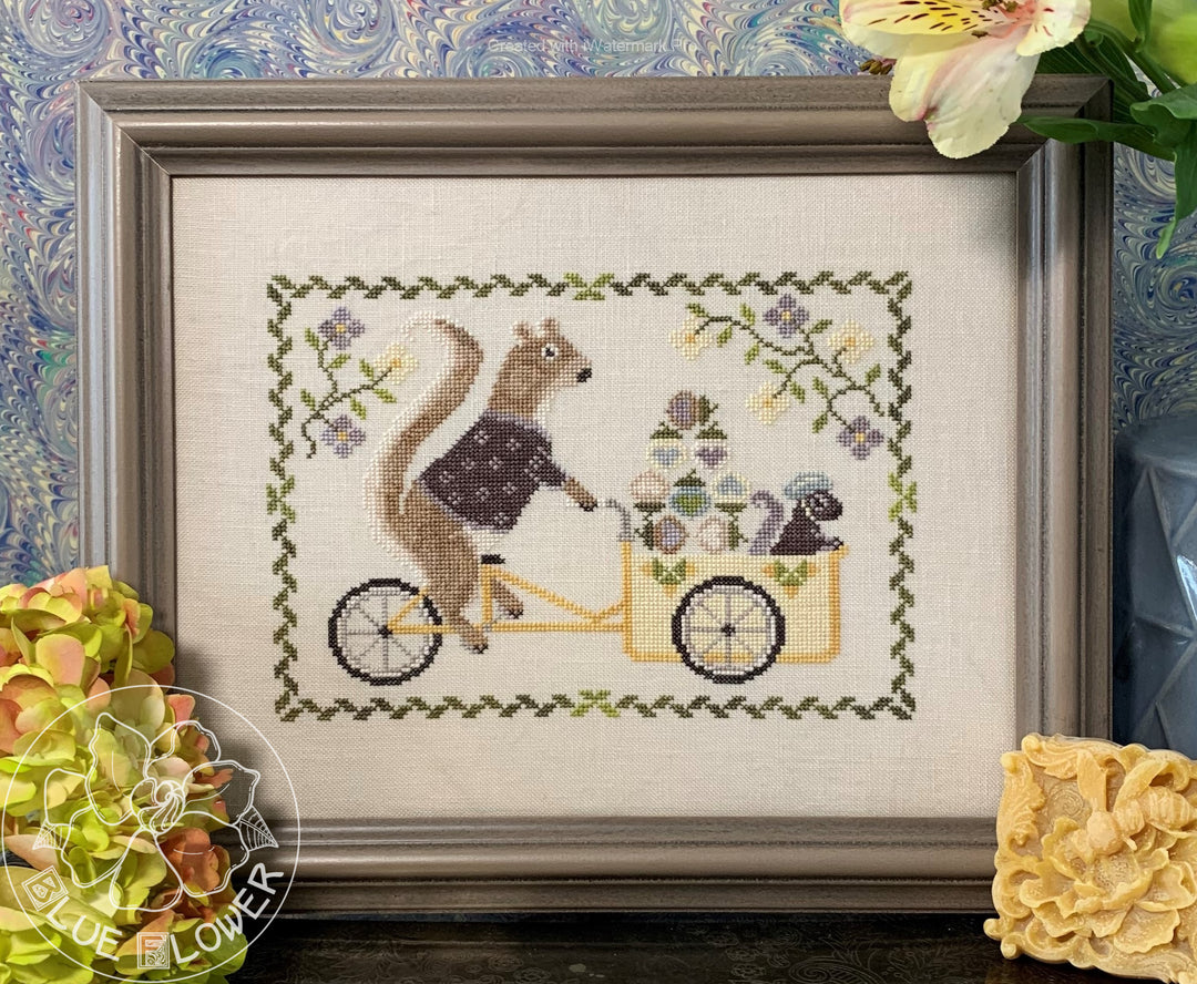 Cross stitch depicting a squirrel with a floral background, framed in a decorative frame, with a small inset showing the texture of the fabric.