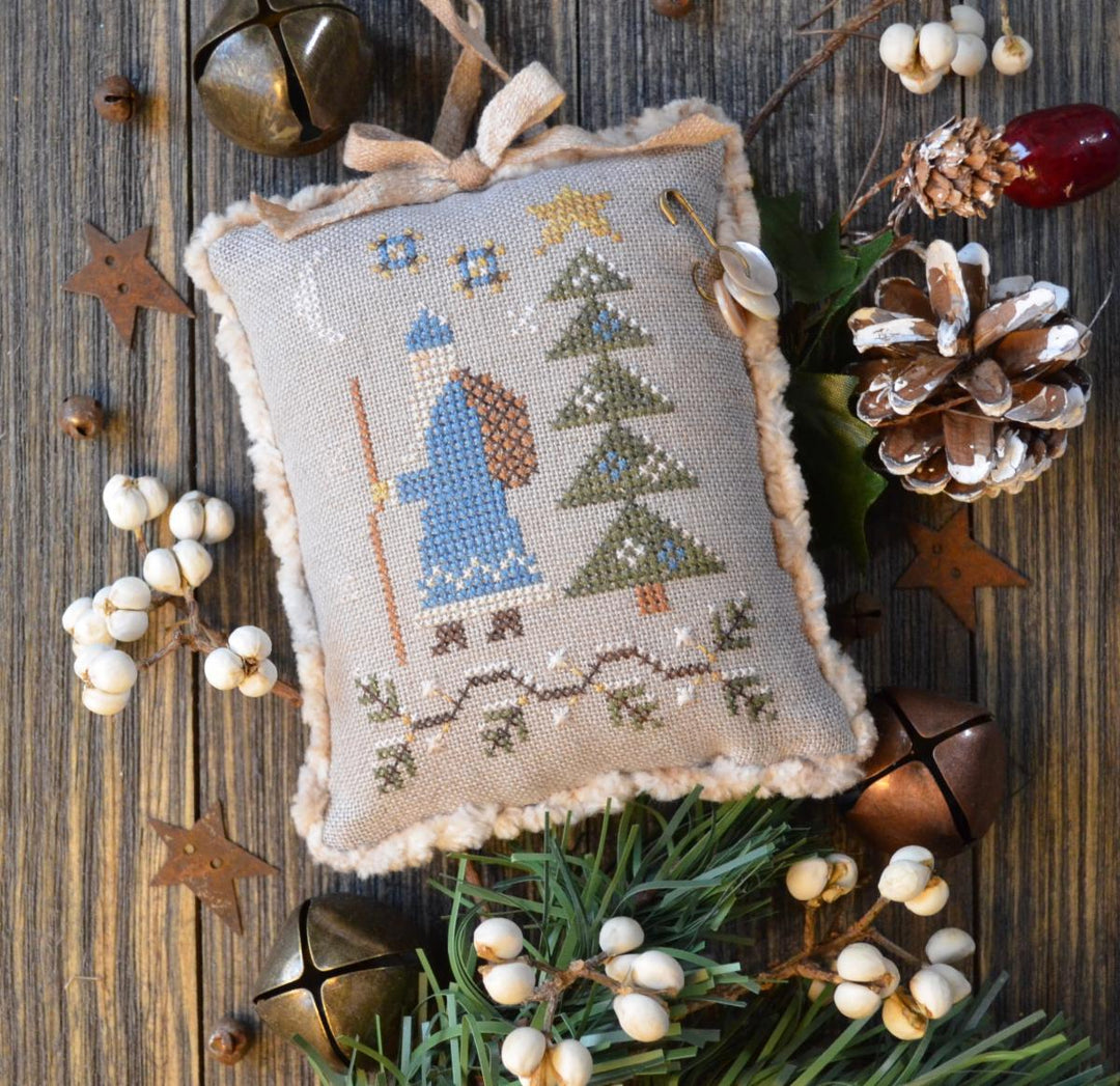 A needlepoint cushion depicting a blue Santa figure with a Christmas tree and decorations, surrounded by a beige border, placed on a wooden surface with holiday-themed decorations around it.