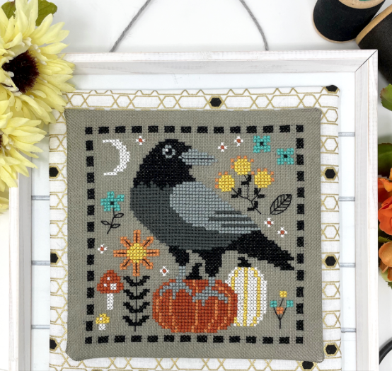 Cross-stitched picture of a crow with pumpkins and flowers on a gray background, framed with decorative elements.