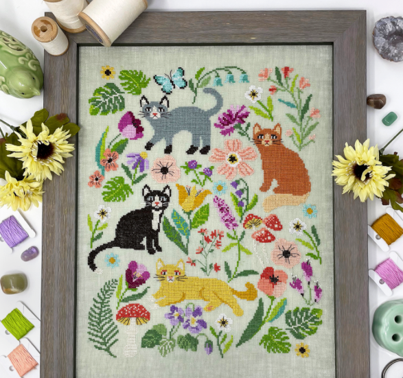 Framed cross-stitched picture of cats and flowers on a textured fabric background.