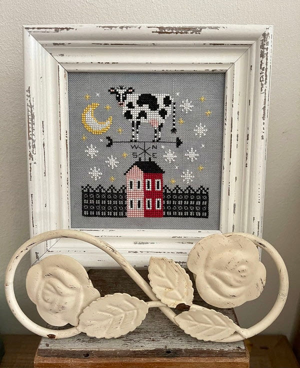 A needlecraft kit depicting a cow with a moon and stars design, stitched on linen, framed in a white wooden frame with a white sash.