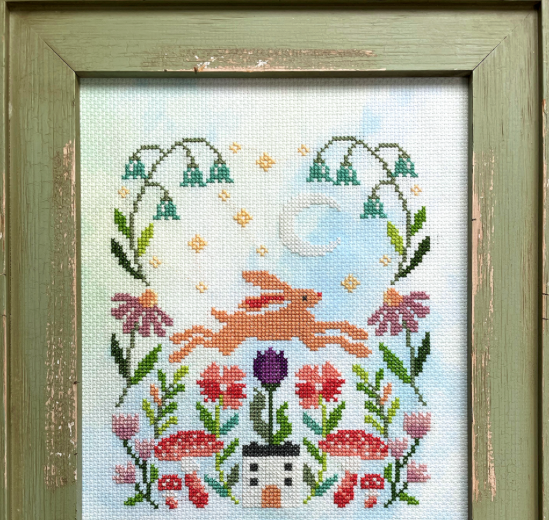 Cross-stitched design of a rabbit among flowers and plants in a wooden frame.