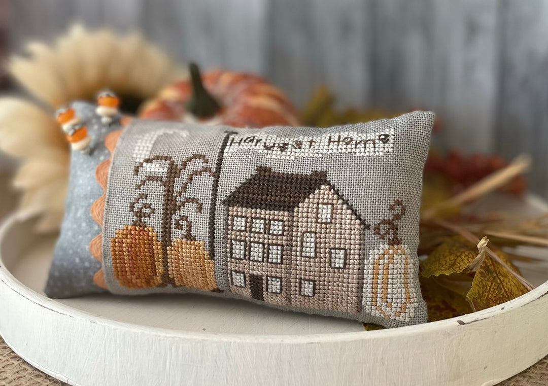 Decorative pillow with cross-stitch design of a house and pumpkins on a wooden surface.