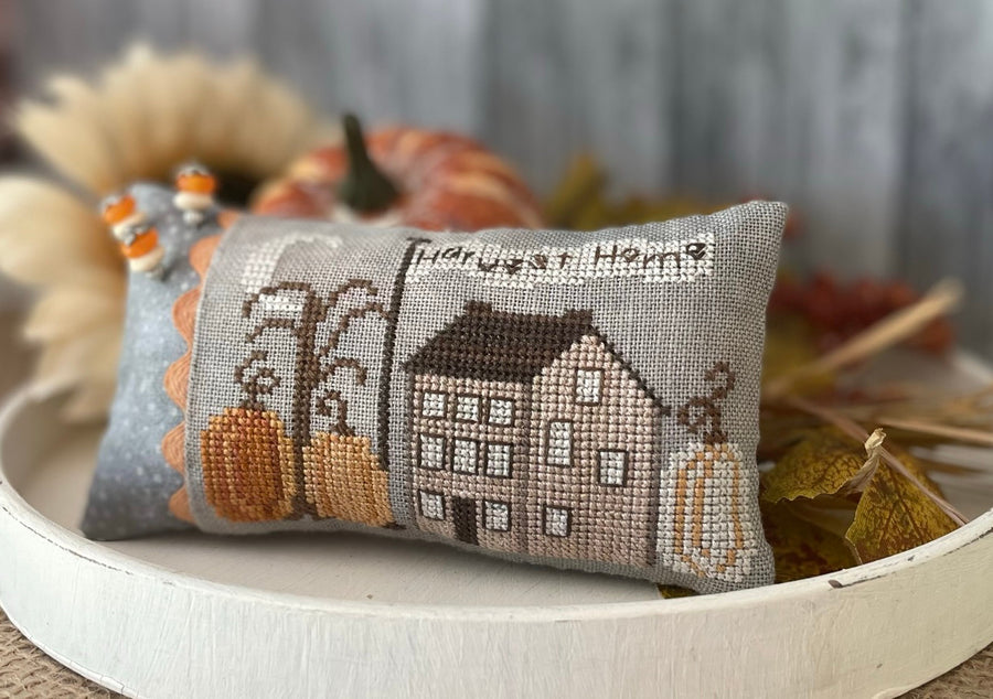 Decorative pillow with cross-stitch design of a house and pumpkins on a wooden surface.