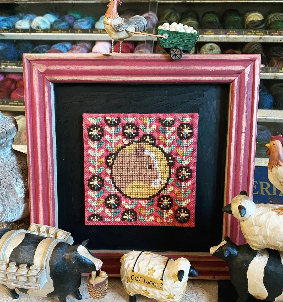 Framed cross-stitched picture of a rooster with yarn and knitting supplies in the background