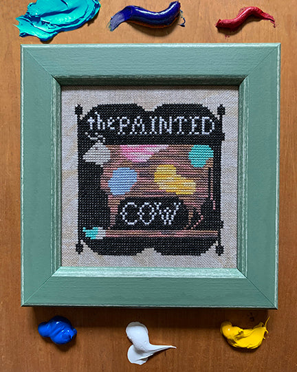 Framed cross-stitched art with 'The Painted Cow' on a wooden surface with fabric swatches.