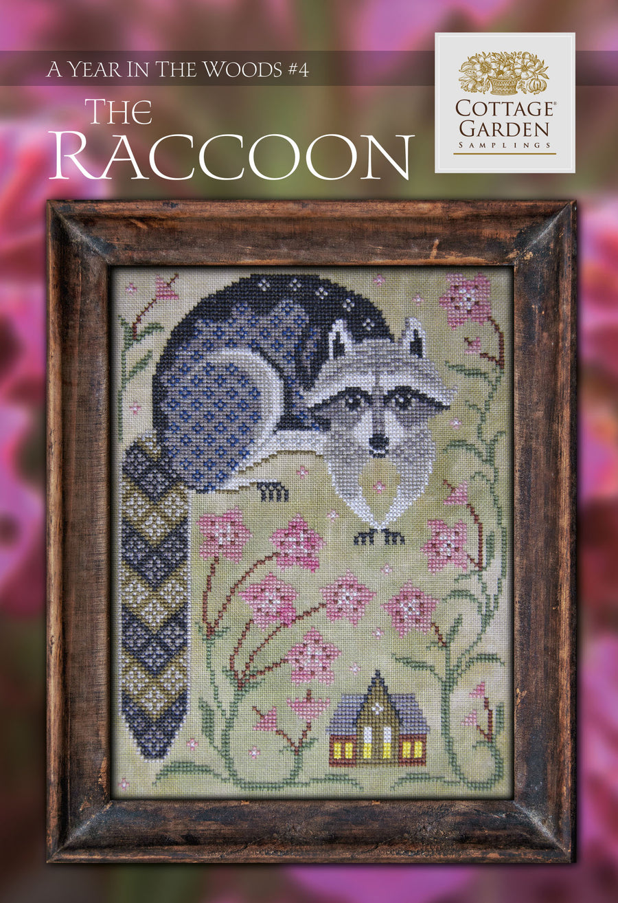 Cross stitch kit depicting a raccoon among flowers with a house in the background, framed in a brown frame.