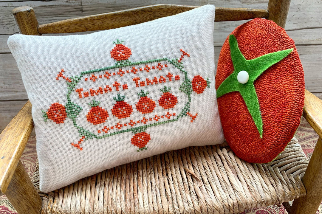 Decorative pillow with cross-stitched design and a tomato-shaped plush toy on a wooden chair.