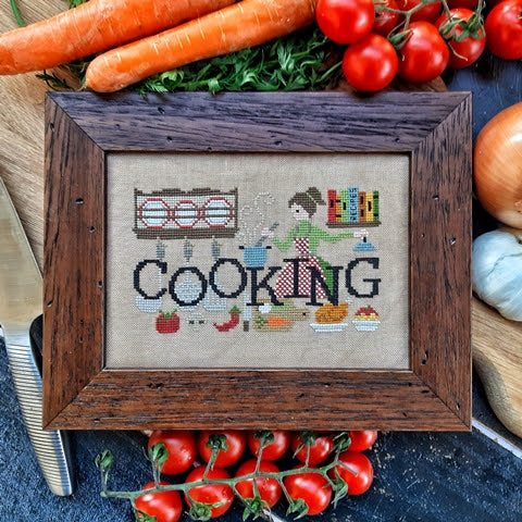 A cross-stitch picture with the word 'COOKING' and kitchen-themed decorations, framed and displayed among vegetables and kitchen items.
