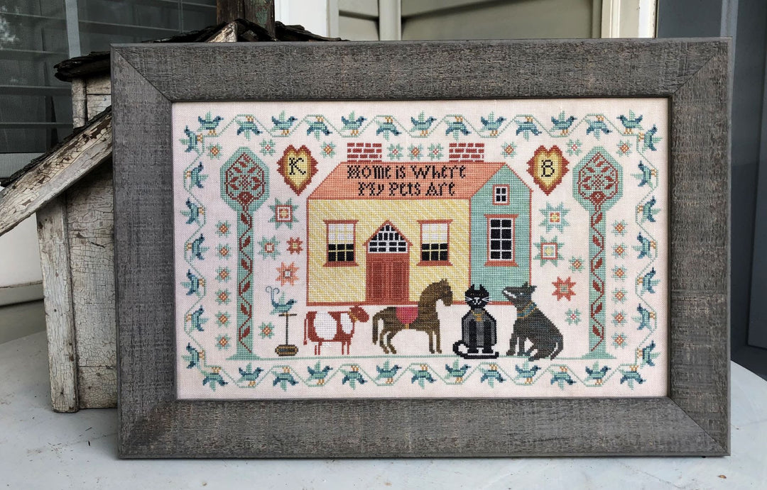 Framed cross-stitched piece with a house and animals on a white background