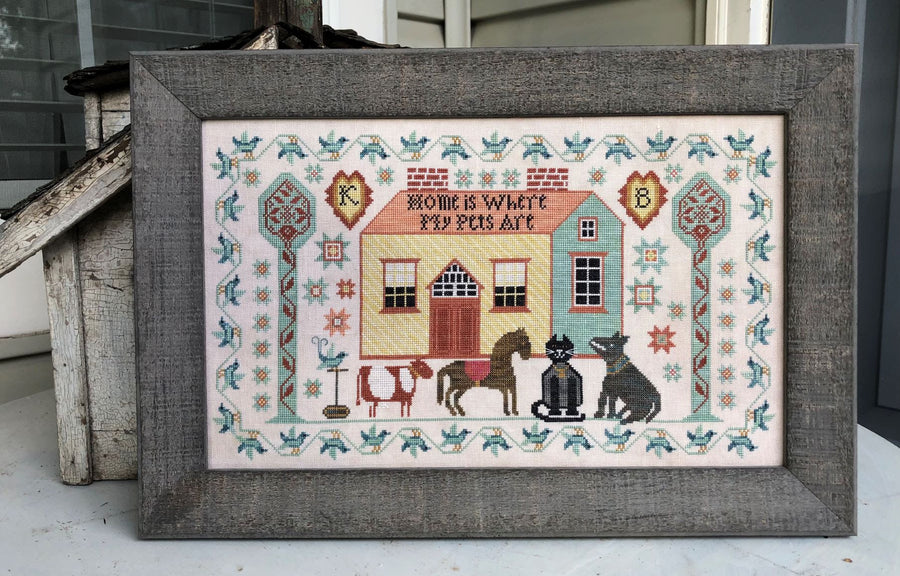 Framed cross-stitched piece with a house and animals on a white background