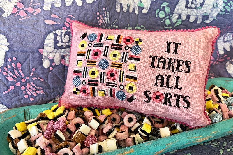 Pink pillow with colorful geometric pattern and 'It takes all sorts' text on a bed with candy.