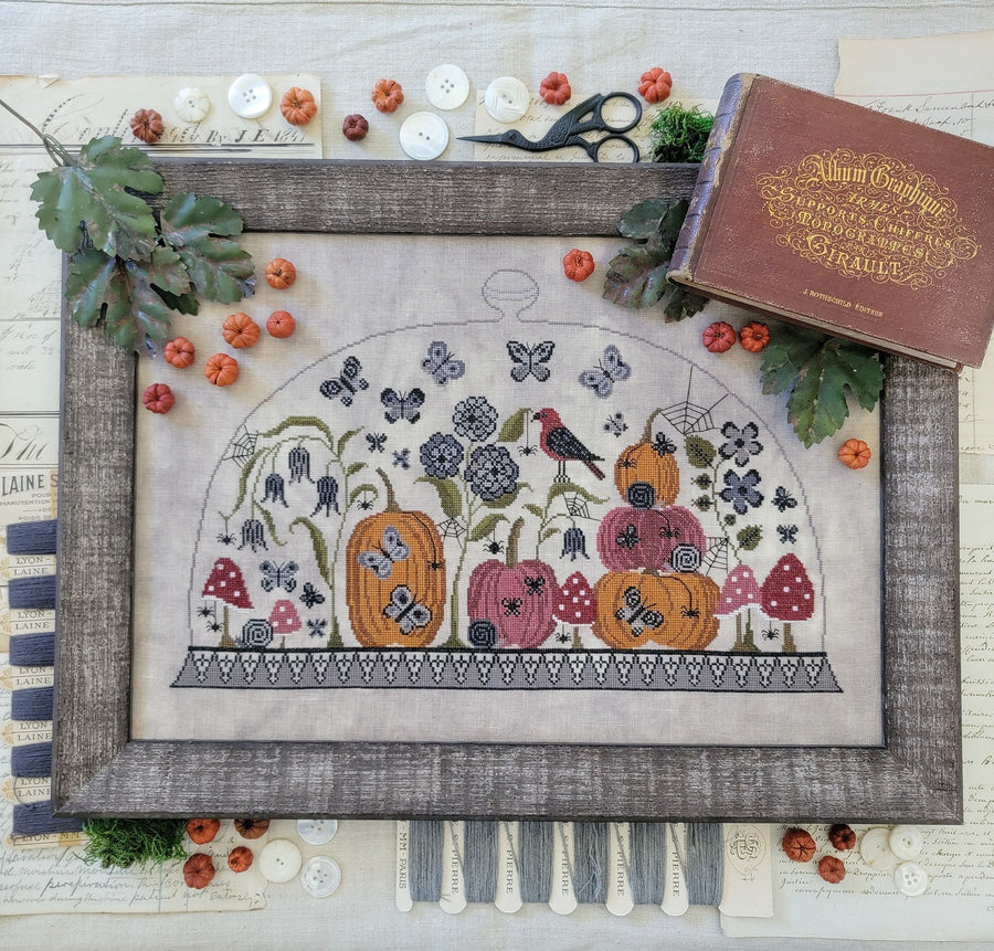 Framed cross-stitched piece with pumpkins, flowers, and a bird on a textured surface.
