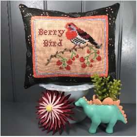 Decorative pillow with cross-stitch design of a bird and berries, placed on a surface with a plant and dinosaur toy.
