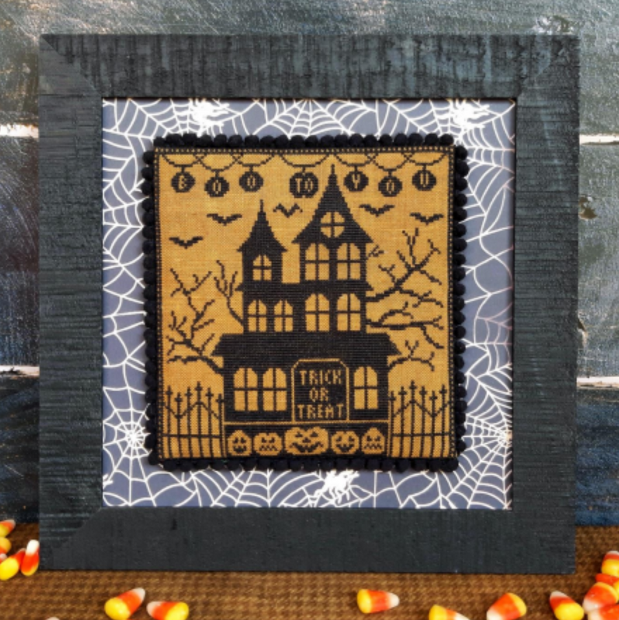 A Halloween-themed cross stitch kit depicting a spooky house with 'TRICK OR TREAT' text, surrounded by bats and pumpkins, encased in a black frame with a spider web pattern.