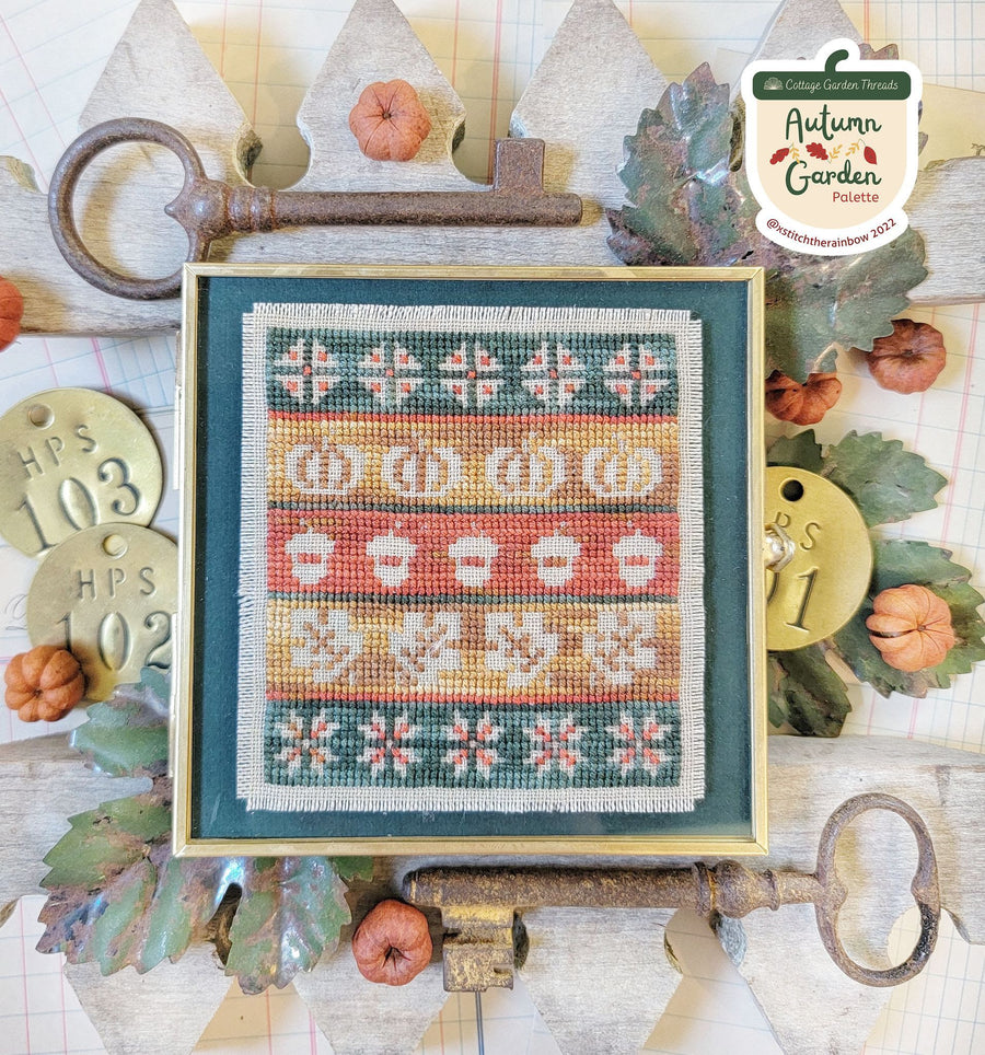 Framed cross-stitched pattern with autumn-themed elements on a decorative background with keys and leaves.