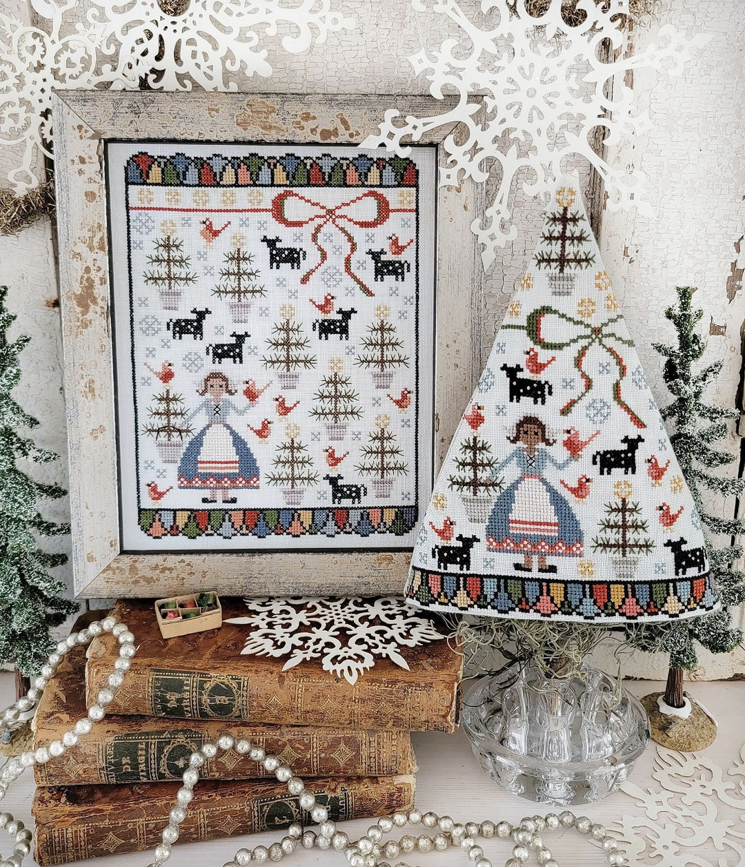 Framed cross-stitched design of a woman with animals and trees, displayed on books with decorative elements.