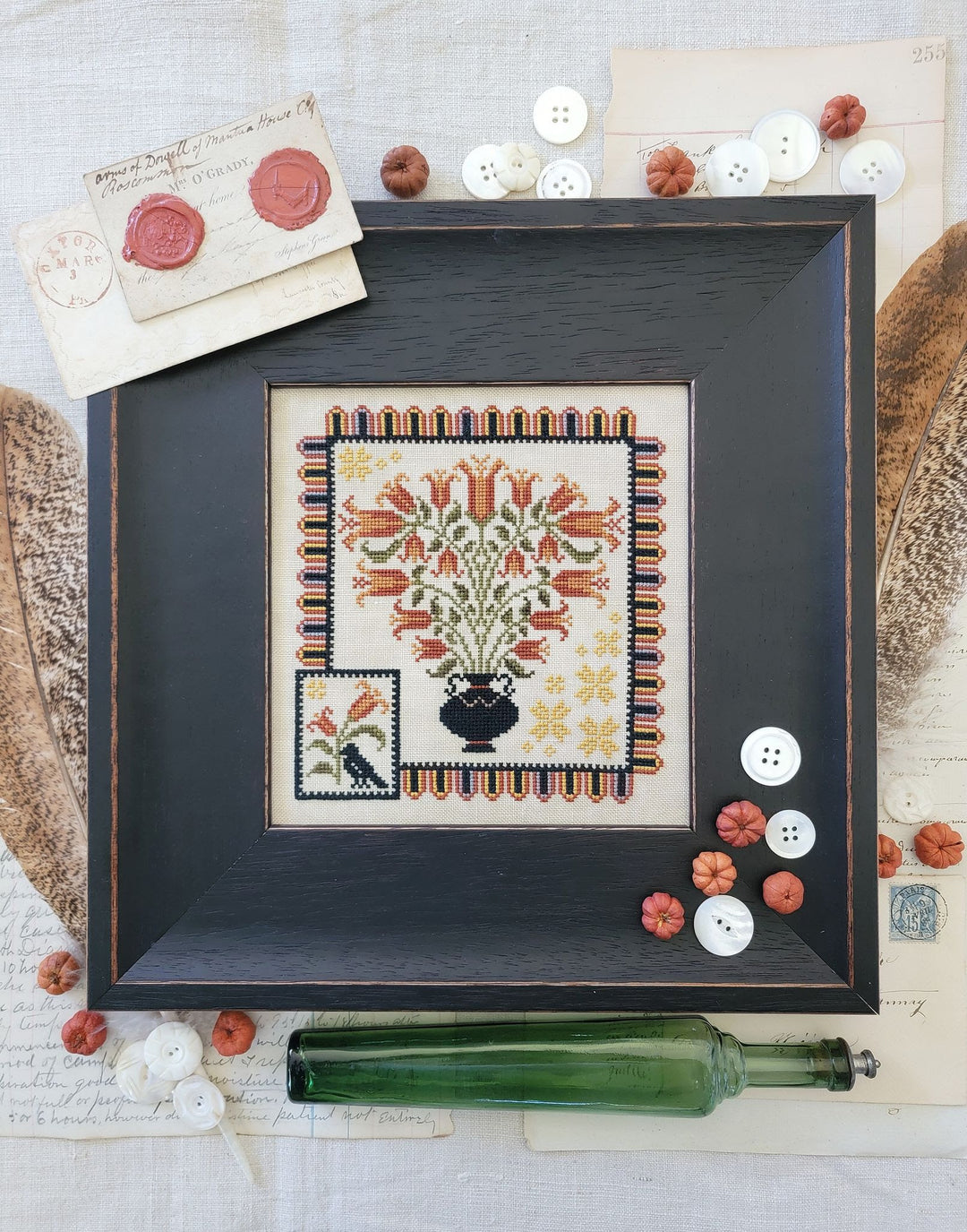 Framed needlepoint with floral design on a textured surface with buttons and a bottle.
