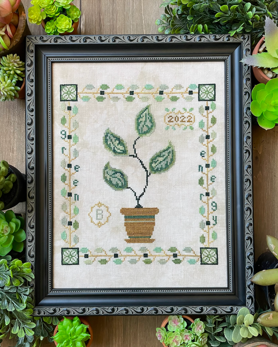 A framed cross-stitch needlework depicting a potted plant with the text '2022' included, set against a wooden background with various greenery.
