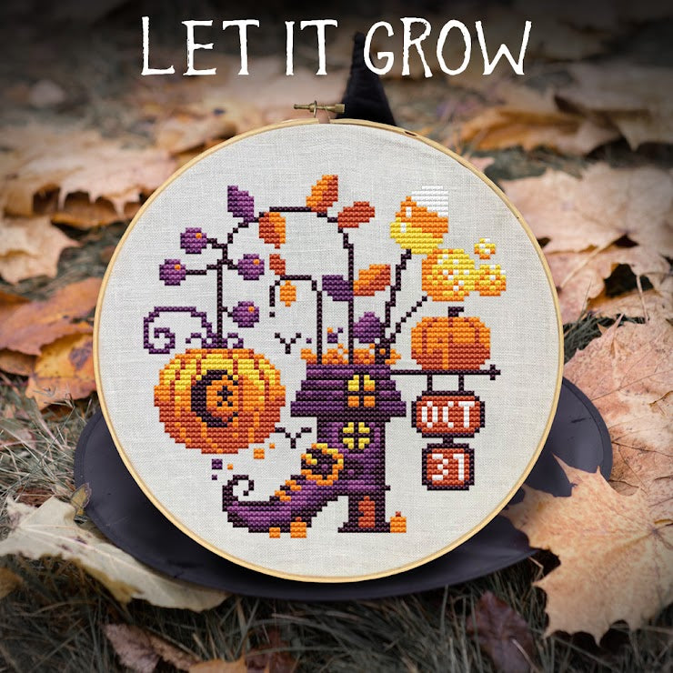 Cross-stitched design of a house with pumpkins and leaves on a fabric hoop, surrounded by autumn leaves.