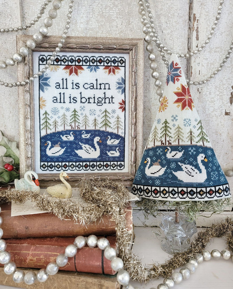 Decorative scene with a framed cross-stitch pattern, swan lamp, and Christmas decorations.
